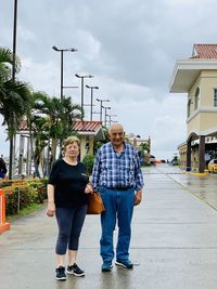 Full length portrait of senior couple standing on footpath against cloudy sky