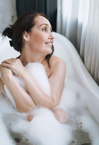 Self loving adult smiling woman with brunette hair taking bath with foam at home