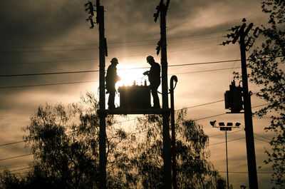 Low angle view of silhouette workers standing on electricity transformer during sunset