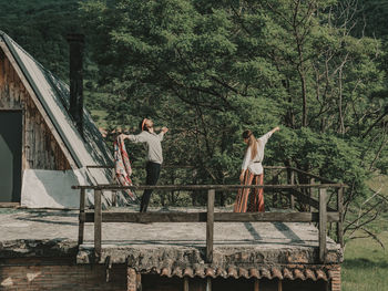 Full body of hippies in boho styled clothes dancing while standing on terrace near wooden cabin during trip in nature