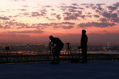 Silhouette man photographing illuminated city against sky during sunset