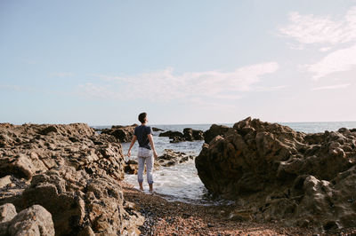 Rear view of young man standing at rocky shore against sky