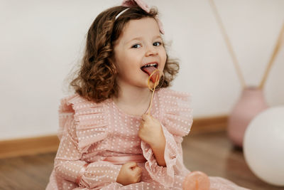 A little girl in a pink dress is sitting on the floor and eating a lollipop