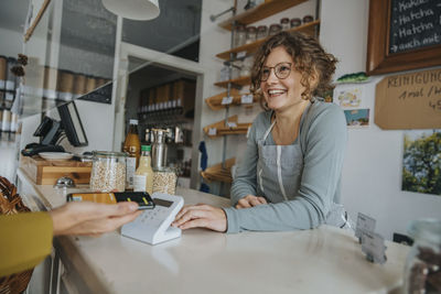 Smiling female owner looking at customer while making payment through smart phone in zero waste shop