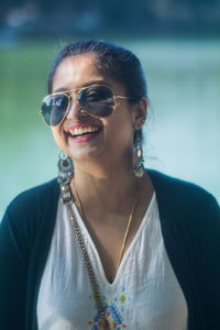 Close-up portrait of cheerful young woman wearing sunglasses while standing against lake