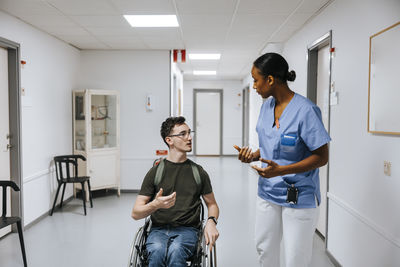 Female doctor discussing with teenage boy sitting on wheelchair at corridor of hospital