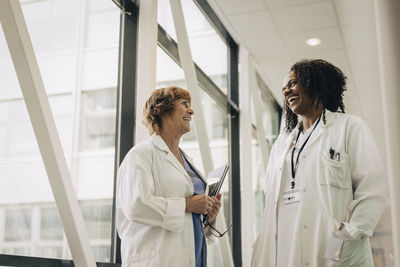 Low angle view of happy female physician with hands in pockets discussing with colleague at hospital