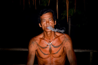 Portrait of shirtless man with tattoo smoking at night
