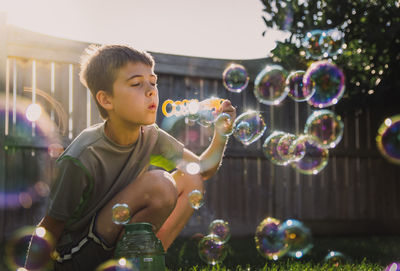 Boy blowing bubbles while crouching at backyard during sunny day