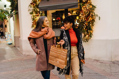 Satisfied multiracial female friends in warm clothes looking at each other while walking on city street after shopping