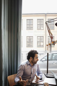 Businessman gesturing while sitting at table against window in creative office