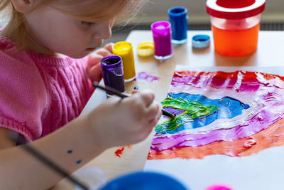 Little girl painting rainbow on paper with colorful paints sitting at the table at home