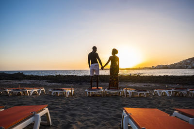 Rear view of man holding hands of woman while standing on sand against sea during sunset