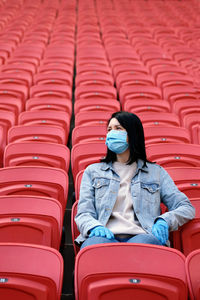 A female fan in a medical mask and rubber gloves sits alone in an empty stadium with red seats.