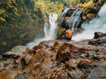 Rear view of man sitting on cliff against waterfall