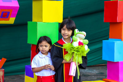 Portrait of girl wearing graduation gown while standing with female friend against colorful toy blocks