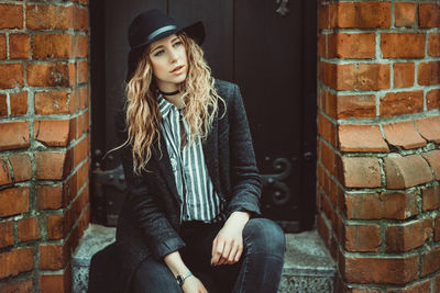 Young woman wearing hat sitting against brick wall 