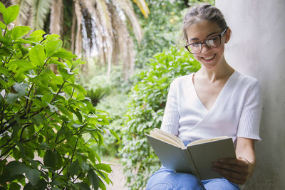 Smiling young woman reading book while sitting by wall