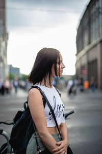 Young woman looking away while standing on city street against sky
