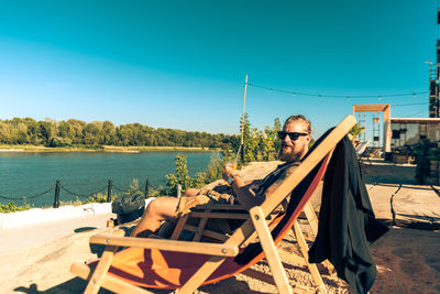 Hipster sitting on folding chair at riverbank against clear blue sky