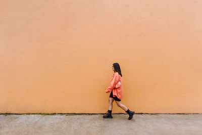 Full body side view of stylish female in trendy outfit and sunglasses walking past orange wall