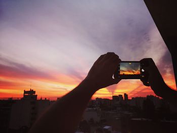 Cropped image of hand holding cityscape against sky during sunset