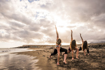 Young women practicing yoga at beach against cloudy sky