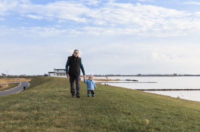 Full length of grandfather and granddaughter holding hands while walking on field by river against sky