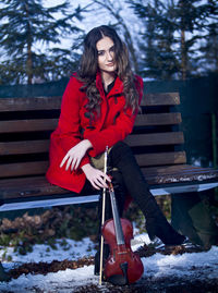 Full length portrait of beautiful woman sitting on bench with violin during winter