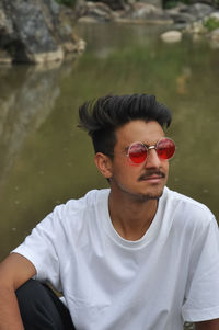 A young guy sitting beside of river with wearing white t-shirt and sunglasses with looking sideways 