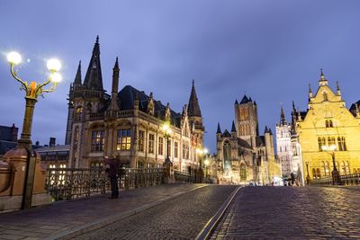 Illuminated street amidst buildings against sky at dusk