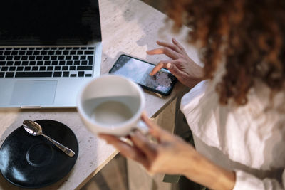 Hands of businesswoman holding coffee cup using smart phone sitting with laptop at desk