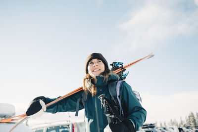 Smiling woman carrying skis during winter against sky