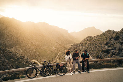 Male and female explorers with bicycles talking while sitting on railing at sunset