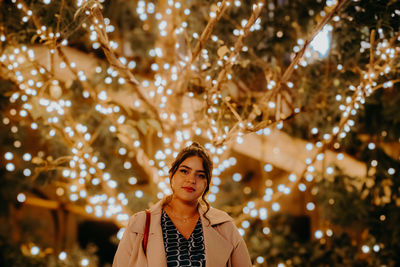 Portrait of woman standing against illuminated christmas tree