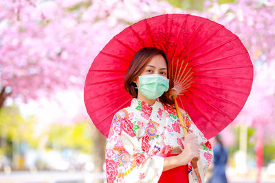 Portrait of woman wearing mask while holding pink umbrella against trees