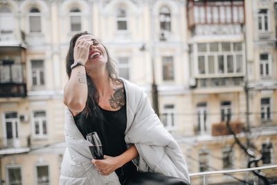 Cheerful woman having red wine while wrapped in blanket at balcony