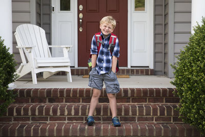 Bashful, smiling, blonde boy stands on steps with hands in pockets