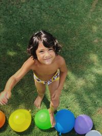 High angle portrait of smiling boy standing by water bomb on grass at back yard