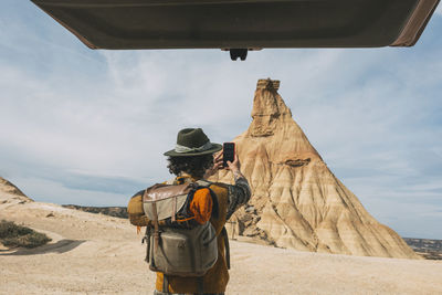 Rear view of a man with a hat taking a photo with a smart phone from his van in the bardenas desert.