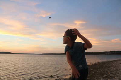 Boy throwing rock in lake against sky during sunset