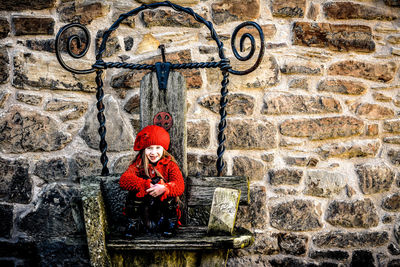 Smiling girl sitting on bench against stone wall during sunny day