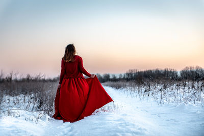 Rear view of woman wearing red dress walking on snowy field during sunset