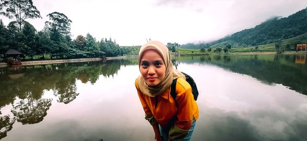 Portrait of smiling young woman standing by lake against trees