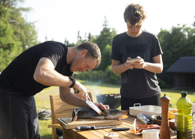 Mature man cutting fish over table while teenage boy using phone in yard