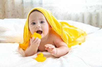 Portrait of cute baby girl holding rubber duck lying with towel on head at home