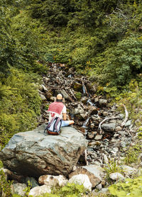 Young man with backpack sitting on large stone looking at mountain stream among forest in hike