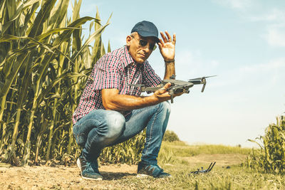 Low angle view of mature man wearing sunglasses holding drone while crouching on field against sky