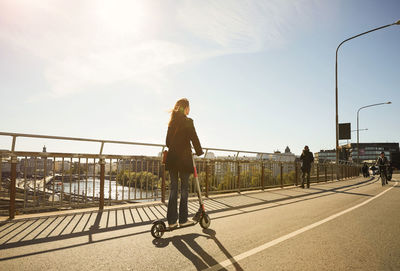 Full length of woman riding electric push scooter on bridge against blue sky