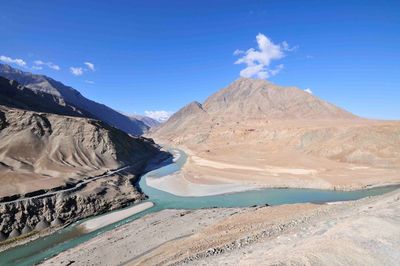 Scenic view of confluence of indus and janskar river and snowcapped mountains against blue sky.